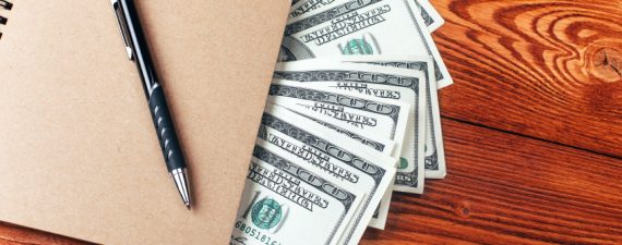 Brown notebook with dollars and pen lying on wooden table with place for text