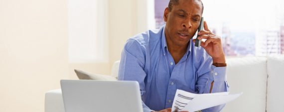 Image of mature man holding papers and talking on phone while sitting at home with a laptop in front