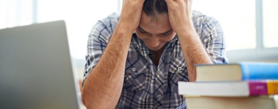 A student sitting with his head in his hand next to a pile of textbooks and his laptop